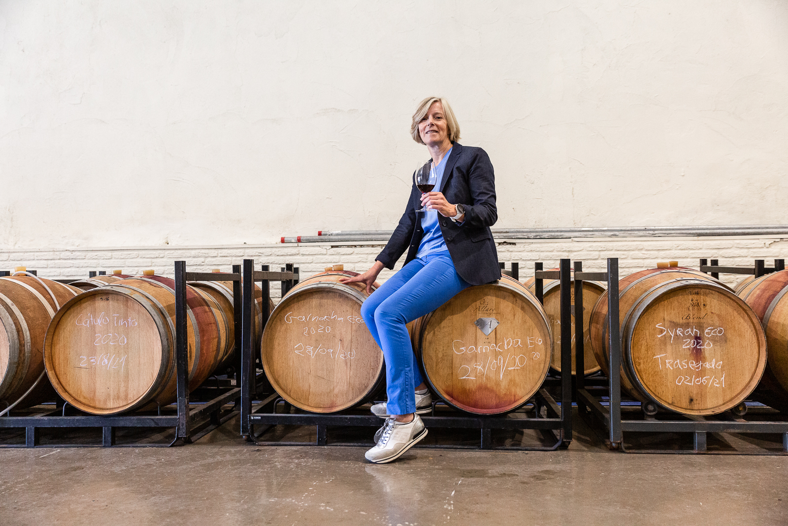 Eugenie, CEO and founder of Axial Vinos, seated on a wine barrel holding a glass of wine